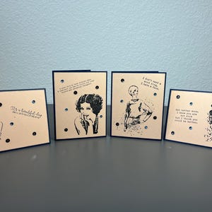 May include: Four greeting cards with black and white illustrations of women and text. The cards have a beige background and blue borders. Each card has small silver embellishments.