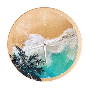 Op de afbeelding: Een ronde houten klok met een strandscène. De wijzerplaat van de klok toont een blauwe oceaan met witte golven die breken op een zandstrand. Op de voorgrond staat een palmboom. De wijzerplaat van de klok heeft Romeinse cijfers en de woorden "TWELVE", "THREE", "SIX" en "NINE".