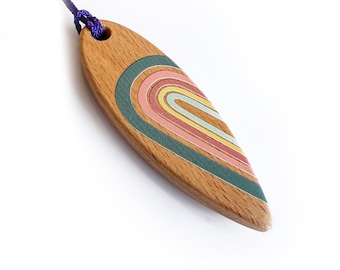 Könnte beinhalten: Holzanhänger in Surfbrettform. Der Anhänger hat ein Regenbogen-Design in Pastellfarben, darunter Rosa, Gelb und Blau, vor hellem braunen Holzmaserung. Eine lila Kordel ist zum Tragen befestigt.