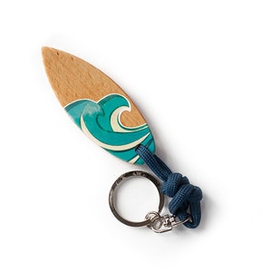 Könnte beinhalten: Ein Holz-Surfbrett-Schlüsselanhänger mit einem blauen und weißen Wellendesign. Der Schlüsselanhänger hat eine dunkelblaue Schnur und einen silbernen Metallring.