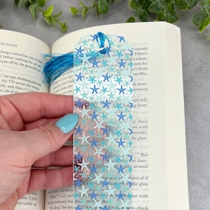 May include: A clear bookmark with a blue tassel and a design of white and blue stars on a light blue background. The bookmark is being held in a hand.