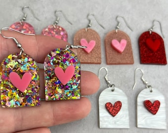 May include: A collection of heart-shaped earrings in various styles. Some earrings feature a pink heart on a glittery background, while others have a solid color or glitter finish. The earrings are designed with a rounded top and a hook for pierced ears.