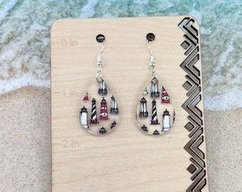 May include: A pair of teardrop-shaped earrings with a white background and a pattern of red and black lighthouses. The earrings are hanging from a wooden display card with a ruler.