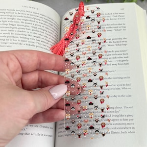 May include: A clear bookmark with a red tassel features a repeating pattern of adorable dog faces in various poses and wearing festive attire. The bookmark is decorated with snowflakes and glitter.