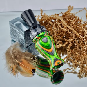 May include: A duck call with a black mouthpiece and a body featuring green, orange, and black wood grain. The call is labeled "High Tide" in white text. A feather and a decorative block are also present.