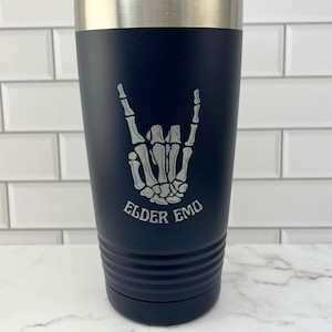 Pode incluir: Um copo de aço inoxidável preto com tampa prateada. O copo tem uma mão de esqueleto branca fazendo um sinal de rock and roll com o texto "Elder Emo" embaixo.