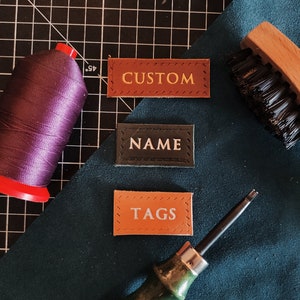 Op de afbeelding: Drie leren labels met gestikte randen. De labels zijn bruin, zwart en oranje. De labels hebben de woorden "CUSTOM", "NAME" en "TAGS" in wit erop.