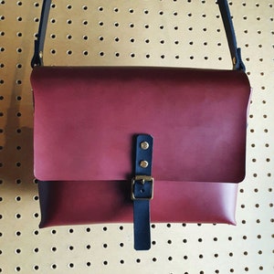 May include: A burgundy leather messenger bag with a black adjustable strap and a brass buckle. The bag has a flap closure and a small pocket on the front.