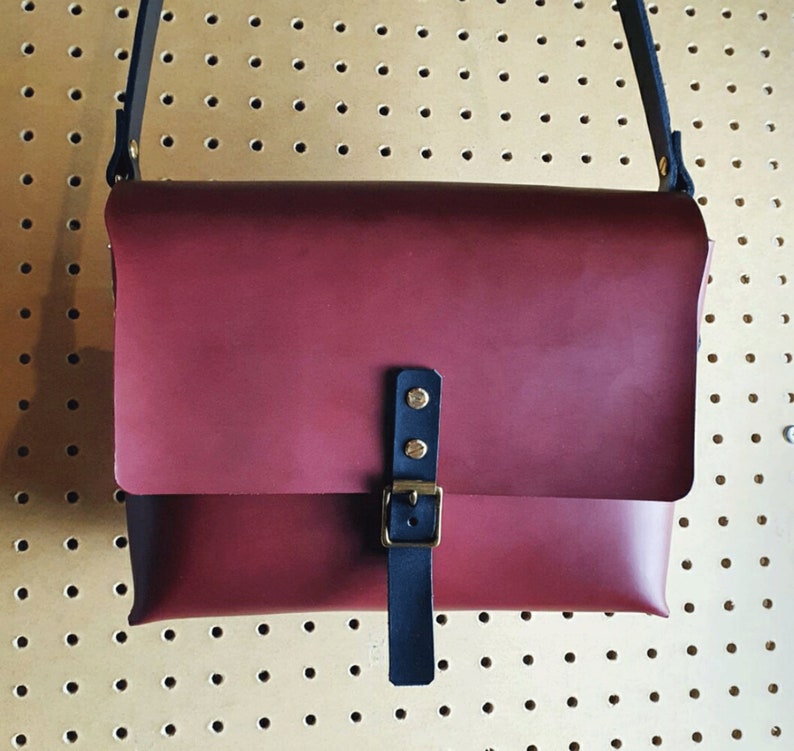 May include: A burgundy leather messenger bag with a black adjustable strap and a brass buckle. The bag has a flap closure and a small pocket on the front.