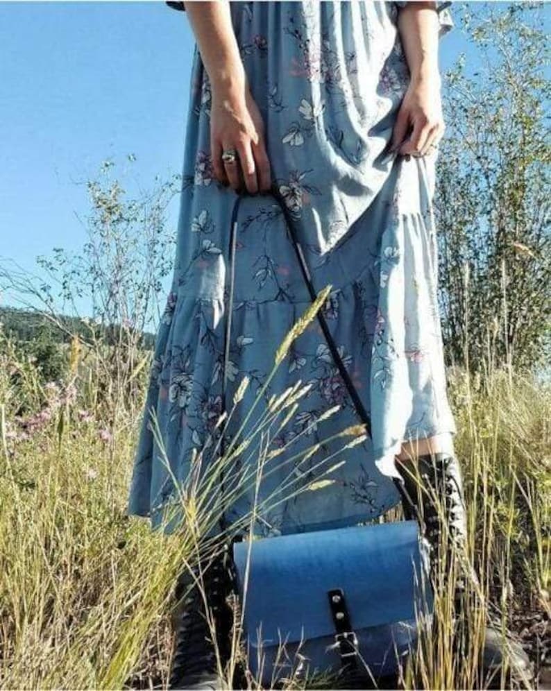 May include: A blue floral dress with a blue leather crossbody bag. The bag has a flap closure with a buckle and an adjustable strap.