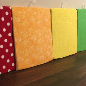 Puede incluir: Una fila de cuadrados de tela coloridos, cada uno de un color diferente del arco&iacute;ris. Las telas incluyen rojo con lunares blancos, naranja, amarillo con lunares blancos, verde, azul claro y morado. Los cuadrados cuelgan de ganchos blancos.
