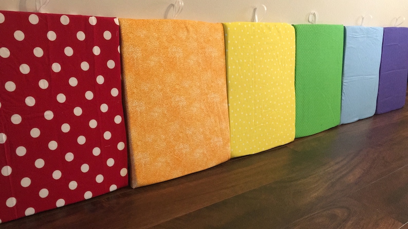 Puede incluir: Una fila de cuadrados de tela coloridos, cada uno de un color diferente del arco&iacute;ris. Las telas incluyen rojo con lunares blancos, naranja, amarillo con lunares blancos, verde, azul claro y morado. Los cuadrados cuelgan de ganchos blancos.