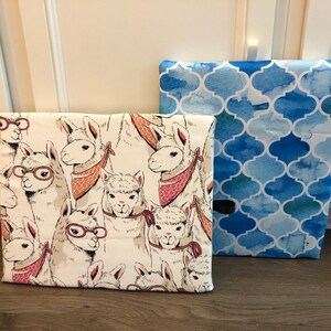 Puede incluir: Dos cajas de almacenamiento de tela. Una presenta un fondo crema con ilustraciones de llamas con gafas y pa&ntilde;uelos. La otra tiene un patr&oacute;n de cuatro hojas de acuarela azul. Ambas cajas son rectangulares y parecen ser de tela.