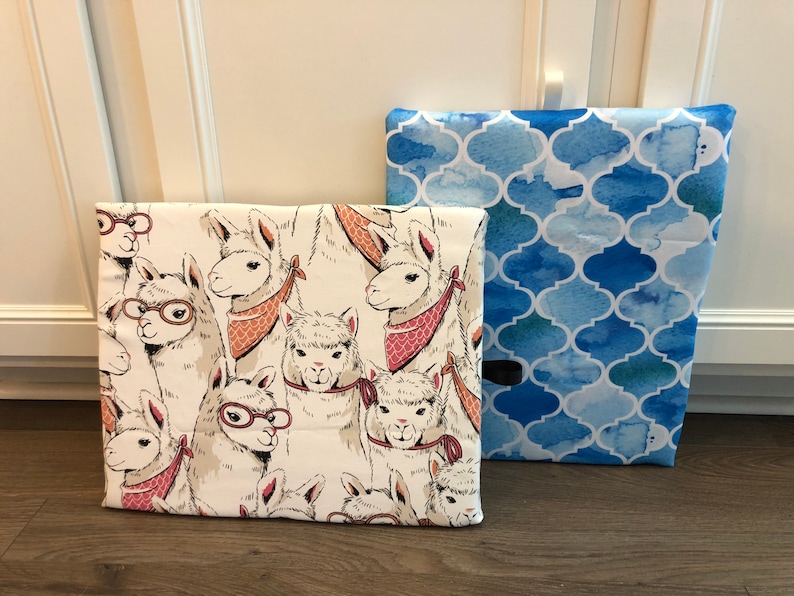 Puede incluir: Dos cajas de almacenamiento de tela. Una presenta un fondo crema con ilustraciones de llamas con gafas y pa&ntilde;uelos. La otra tiene un patr&oacute;n de cuatro hojas de acuarela azul. Ambas cajas son rectangulares y parecen ser de tela.