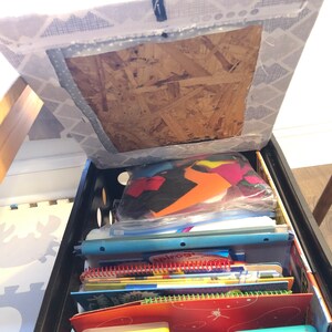 Puede incluir: Una caja de pl&aacute;stico negra llena de &uacute;tiles escolares, incluyendo cuadernos y carpetas. La caja est&aacute; colocada debajo de una silla con un asiento de tela estampada. El contenido est&aacute; bien organizado, sugiriendo una soluci&oacute;n de almacenamiento.