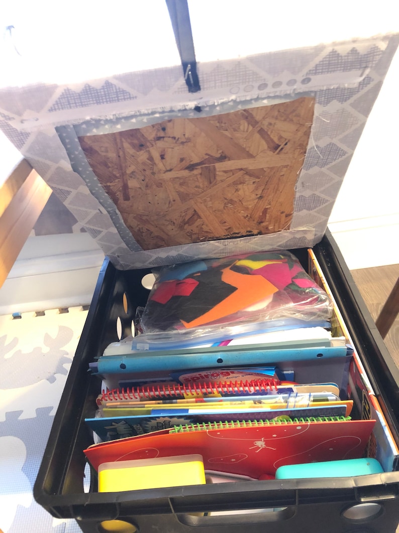 Puede incluir: Una caja de pl&aacute;stico negra llena de &uacute;tiles escolares, incluyendo cuadernos y carpetas. La caja est&aacute; colocada debajo de una silla con un asiento de tela estampada. El contenido est&aacute; bien organizado, sugiriendo una soluci&oacute;n de almacenamiento.