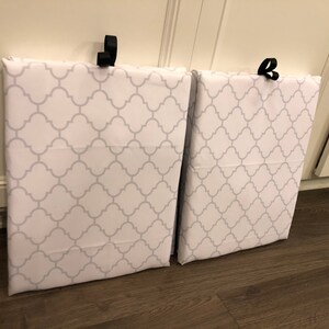 Puede incluir: Dos bolsas de almacenamiento de tela blanca con un patr&oacute;n de cuadrifolio gris. Cada bolsa tiene un lazo negro en la parte superior para colgar. Las bolsas est&aacute;n apoyadas contra un armario blanco.