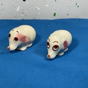 May include: Two ceramic dog figurines. Each dog is white with brown eyes, a pink ear, and a pink spot on its side. The dogs are small and have a whimsical design.