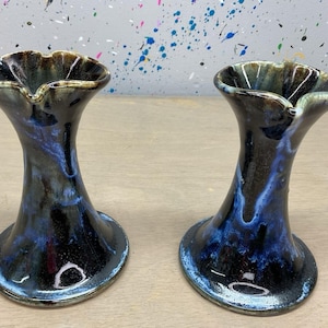 May include: Two handmade ceramic vases with a flared, ruffled rim. The vases have a dark, glossy finish with blue and black color variations. The base is round and the overall shape is elegant and unique.