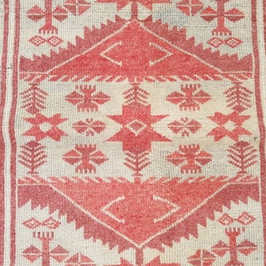 K&ouml;nnte beinhalten: Nahaufnahme eines gewebten Teppichs mit cremefarbenem Hintergrund und einem sich wiederholenden geometrischen Muster in Rott&ouml;nen. Das Design umfasst Rauten, Kreuze und stilisierte florale Elemente, die eine strukturierte, dekorative Oberfl&auml;che schaffen.