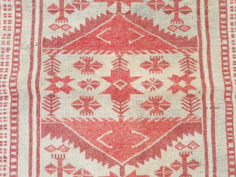 K&ouml;nnte beinhalten: Nahaufnahme eines gewebten Teppichs mit cremefarbenem Hintergrund und einem sich wiederholenden geometrischen Muster in Rott&ouml;nen. Das Design umfasst Rauten, Kreuze und stilisierte florale Elemente, die eine strukturierte, dekorative Oberfl&auml;che schaffen.