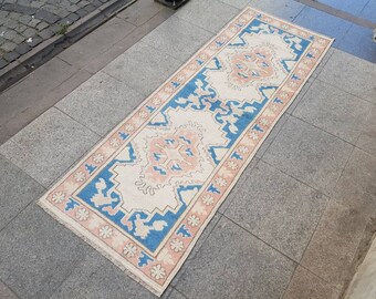 Puede incluir: Una alfombra de pasillo vintage con fondo crema y un patrón geométrico azul y rosa. La alfombra tiene un borde con flecos.