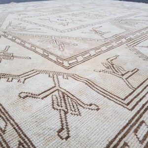 Puede incluir: Una alfombra blanca con un patr&oacute;n geom&eacute;trico marr&oacute;n. El patr&oacute;n presenta dise&ntilde;os repetidos de p&aacute;jaros estilizados y formas geom&eacute;tricas.