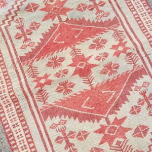 K&ouml;nnte beinhalten: Ein rechteckiger Teppich mit cremefarbenem Hintergrund und einem sich wiederholenden geometrischen Muster in Rot. Das Design umfasst sternartige Formen, Zickzacklinien und kreuzartige Motive, die von einem roten Rand mit einem sich wiederholenden quadratischen Muster umrahmt werden. Der Teppich hat ein Vintage-Aussehen.