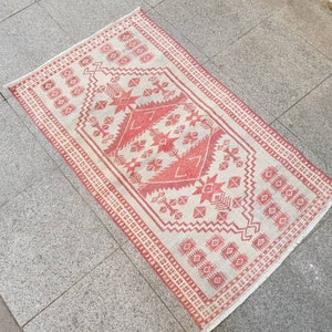 K&ouml;nnte beinhalten: Ein rechteckiger Teppich mit cremefarbenem Hintergrund und rotem geometrischem Muster. Das Muster umfasst sternartige Formen, Rauten und andere geometrische Designs. Der Teppich hat einen roten Rand und liegt auf einer grauen Steinoberfl&auml;che.