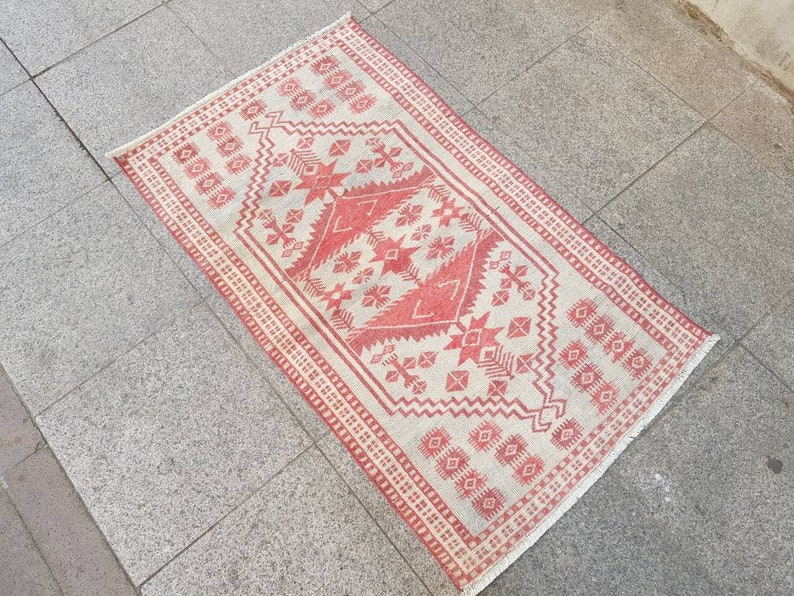 K&ouml;nnte beinhalten: Ein rechteckiger Teppich mit cremefarbenem Hintergrund und rotem geometrischem Muster. Das Muster umfasst sternartige Formen, Rauten und andere geometrische Designs. Der Teppich hat einen roten Rand und liegt auf einer grauen Steinoberfl&auml;che.