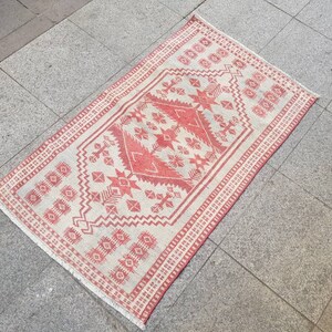 K&ouml;nnte beinhalten: Ein rechteckiger, handgewebter Teppich mit einem geometrischen Muster in Rot und Off-White. Der Teppich hat ein zentrales Design mit sternartigen Formen und einen Rand mit sich wiederholenden Motiven. Der Teppich liegt auf einer grauen Fliesenoberfl&auml;che.