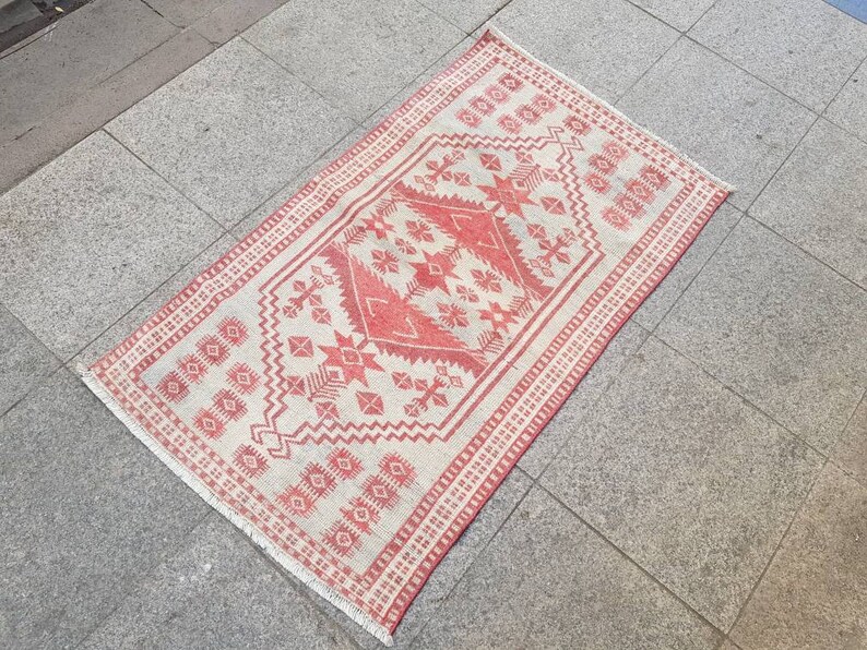 K&ouml;nnte beinhalten: Ein rechteckiger, handgewebter Teppich mit einem geometrischen Muster in Rot und Off-White. Der Teppich hat ein zentrales Design mit sternartigen Formen und einen Rand mit sich wiederholenden Motiven. Der Teppich liegt auf einer grauen Fliesenoberfl&auml;che.