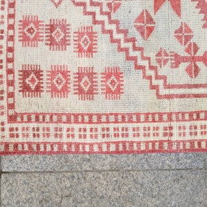 K&ouml;nnte beinhalten: Nahaufnahme eines Teils eines Teppichs mit einem geometrischen Muster in Rot und Off-White. Der Teppich weist sich wiederholende Diamanten- und Rechteckformen mit einem Fransenrand auf. Das Design ist symmetrisch und traditionell, geeignet f&uuml;r die Heimdekoration.