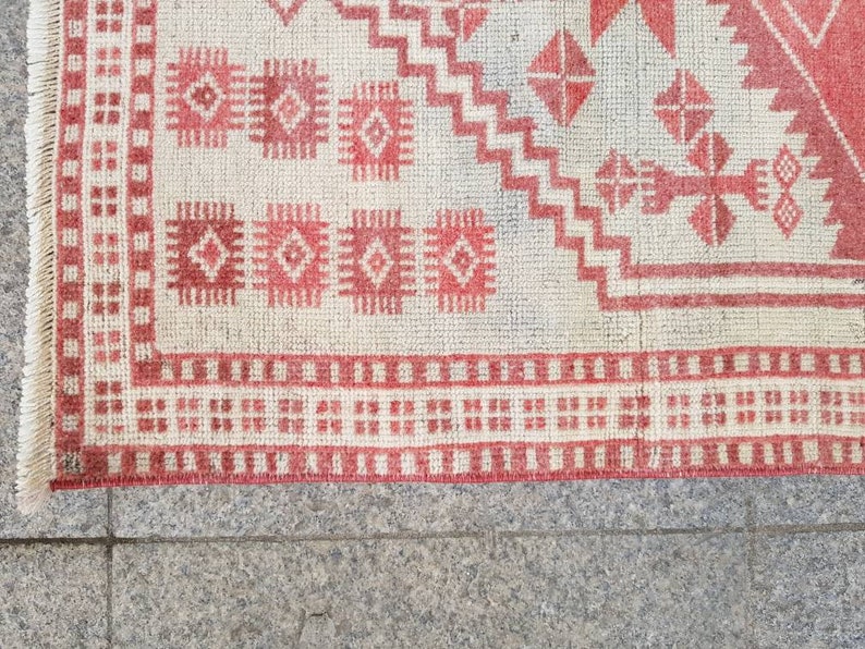 K&ouml;nnte beinhalten: Nahaufnahme eines Teils eines Teppichs mit einem geometrischen Muster in Rot und Off-White. Der Teppich weist sich wiederholende Diamanten- und Rechteckformen mit einem Fransenrand auf. Das Design ist symmetrisch und traditionell, geeignet f&uuml;r die Heimdekoration.