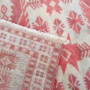 K&ouml;nnte beinhalten: Nahaufnahme eines rot-cremefarbenen Teppichs mit geometrischem Muster. Der Teppich hat einen detaillierten Rand mit Fransen an einer Kante. Das Design zeigt sich wiederholende Blumen- und sternartige Formen in Rott&ouml;nen auf cremefarbenem Hintergrund.