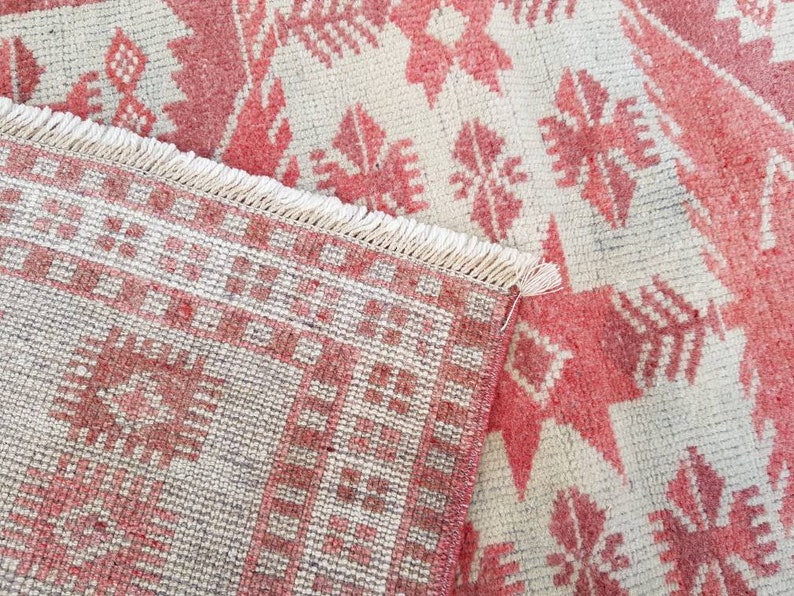 K&ouml;nnte beinhalten: Nahaufnahme eines rot-cremefarbenen Teppichs mit geometrischem Muster. Der Teppich hat einen detaillierten Rand mit Fransen an einer Kante. Das Design zeigt sich wiederholende Blumen- und sternartige Formen in Rott&ouml;nen auf cremefarbenem Hintergrund.