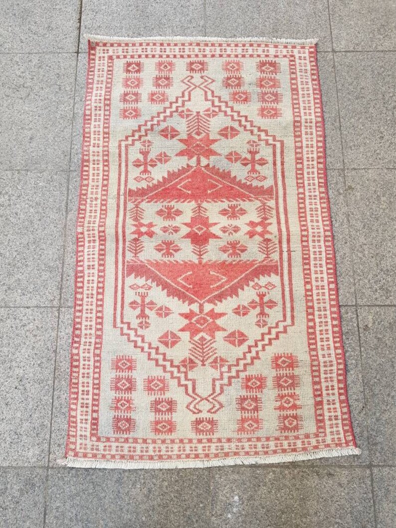 K&ouml;nnte beinhalten: Ein rechteckiger Teppich mit cremefarbenem Hintergrund und rotem geometrischem Muster. Der Teppich hat ein zentrales Design mit Diamanten und Sternen, das von einem roten Rand umrahmt wird. Der Teppich hat an den kurzen Enden einen Fransen.