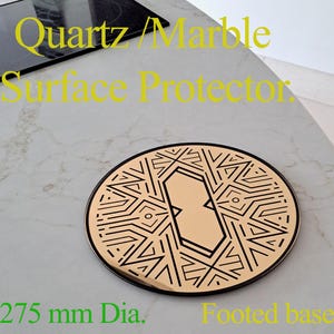 Puede incluir: Un protector de superficie redondo de color dorado con un diseño geométrico negro. El protector tiene una base con pie y mide 275 mm de diámetro.  "Quartz/Marble Surface Protector" está escrito en amarillo en la parte superior de la imagen.