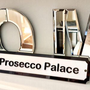 May include: A black and white sign with the text "Prosecco Palace" on a white background. The sign is framed with a black border and has a decorative edge. The sign is hanging on a wall with mirrored letters spelling out "HOME" above it.