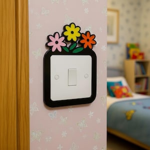 May include: A decorative light switch cover featuring a black frame with colorful flower accents. The flowers are pink, yellow, and orange. The switch plate is white with a single toggle switch. The cover is mounted on a pink wall with a butterfly pattern.