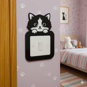 May include: A black and white cat-shaped light switch cover. The cat is peeking over a white light switch plate. The wall is lavender with paw prints and yarn ball decals. A framed cat silhouette art piece is visible in the background.