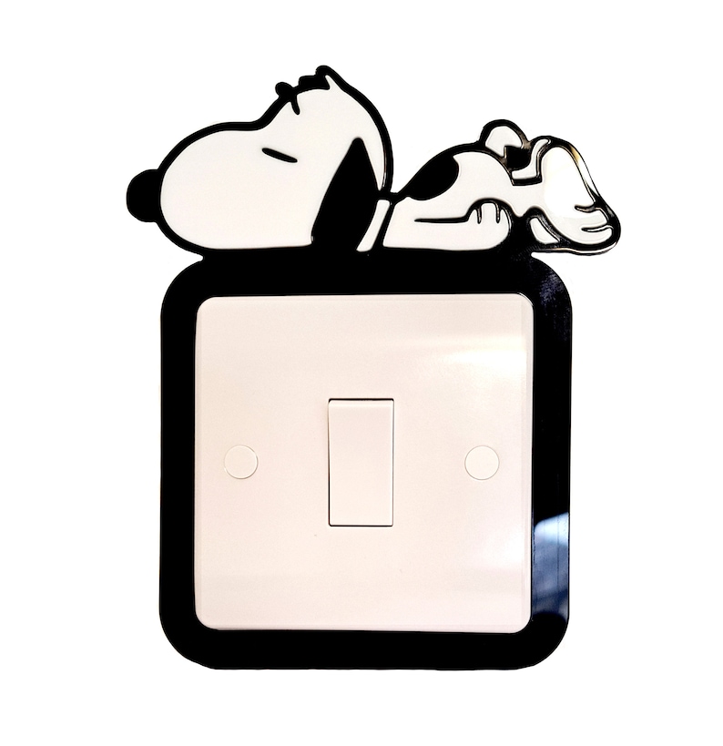 May include: A black and white cartoon image of Snoopy, the beagle from the Peanuts comic strip, lying on a white light switch cover with a black border. The light switch is a standard single-pole switch with a white toggle.