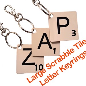 May include: Keyrings featuring Scrabble tiles spelling "ZAP". The tiles are off-white with black letters and numbers, attached to silver keychains. The image includes the text "Large Scrabble Tile Letter Keyrings".