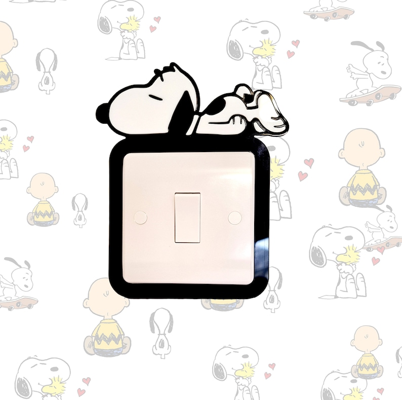 May include: A black and white light switch cover with a cartoon image of Snoopy lying down on top of it. The background is white with a pattern of cartoon Snoopy images.
