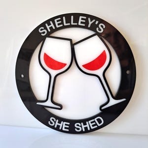 May include: Black and white round sign with the text "Shelley's She Shed" and two red wine glasses clinking together.