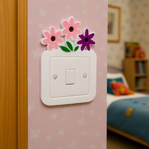 Puede incluir: Una cubierta de interruptor de luz blanca decorada con un diseño floral. Las flores son rosas y moradas con hojas verdes. El interruptor de luz está montado en una pared rosa con un patrón de mariposas.