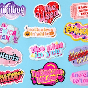 Puede incluir: Una colección de 12 pegatinas coloridas con varios nombres de bandas y títulos de canciones. Las pegatinas son de varias formas y tamaños, incluyendo corazones, nubes y rectángulos. Algunas de las pegatinas tienen un acabado brillante. Las pegatinas están todas sobre un fondo azul claro.