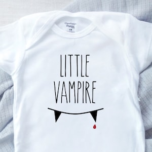 May include: White baby bodysuit featuring the words "LITTLE VAMPIRE" in black, with a fang graphic and a red blood drop. The garment is a Gerber Organic 12M size. A cute Halloween-themed outfit.