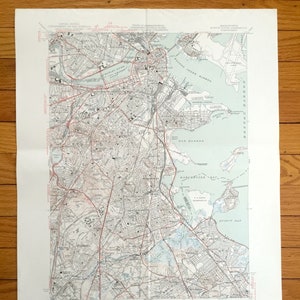 May include: A vintage map of Boston, Massachusetts, showing the city's streets, waterways, and landmarks. The map is printed in black and white with red lines for roads and blue for water. The map includes the Charles River, Boston Harbour, and the surrounding areas.