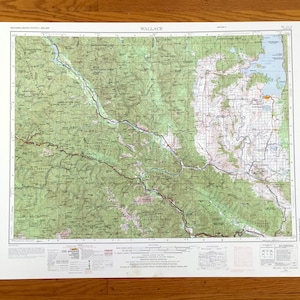 May include: A detailed topographic map titled "Wallace" with green, brown, and blue hues. The map shows a mountainous region with rivers, forests, and elevation contours. The map is printed on white paper and is set against a wooden background.